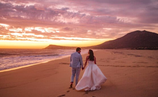 A couple in wedding attire walks hand in hand along a sandy beach at sunset, leaving footprints behind. The sky is filled with dramatic clouds and warm colors, with hills visible in the distance. by Justin Salem Meyer