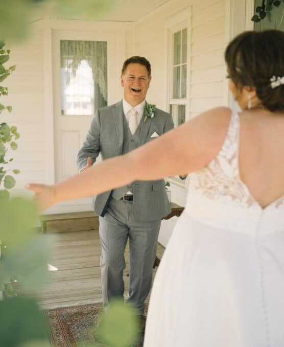 A groom in a light gray suit smiles warmly with open arms as he sees his bride, who is wearing a white dress with lace details, during a joyful first look on a porch. by Justin Salem Meyer