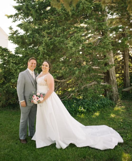 A bride in a white gown holding a bouquet stands next to a groom in a light gray suit. They are outdoors, smiling, with tall green trees in the background. by Justin Salem Meyer
