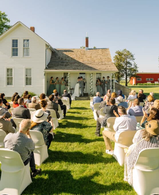 A wedding ceremony takes place outdoors on a sunny day, with guests seated on white-covered chairs facing a couple exchanging vows on the porch of a white house. Bridesmaids and groomsmen stand by the couple. by Justin Salem Meyer