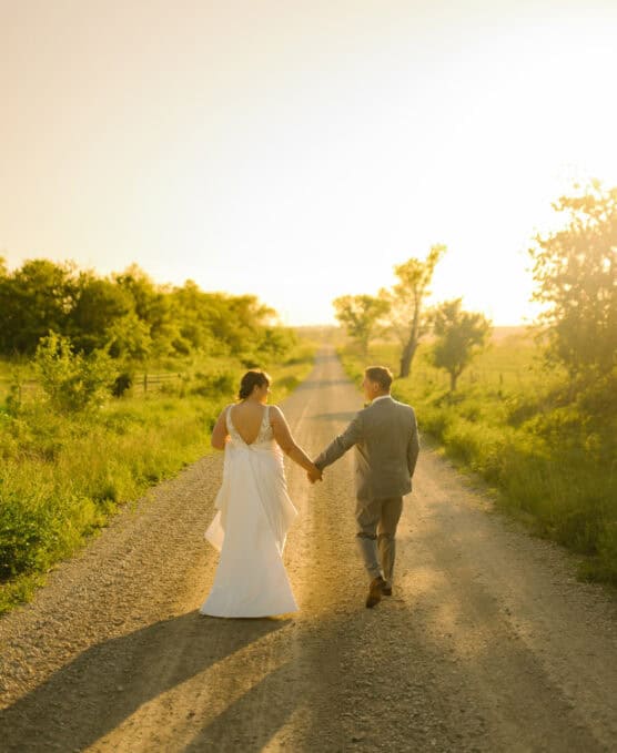 A bride and groom walk hand in hand down a sunlit gravel road surrounded by greenery, with warm golden sunlight casting long shadows behind them. by Justin Salem Meyer