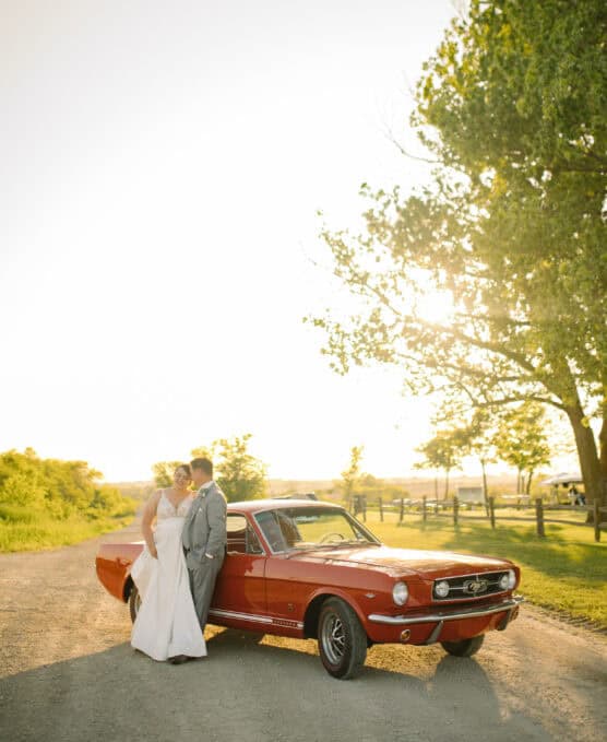 A bride and groom stand close together, smiling beside a classic red Ford Mustang on a sunlit country road, with trees and a wooden fence in the background. by Justin Salem Meyer