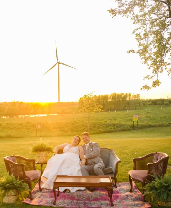A bride and groom sit closely on a vintage sofa outdoors at sunset, surrounded by armchairs and plants, with a wind turbine and green landscape in the background. by Justin Salem Meyer