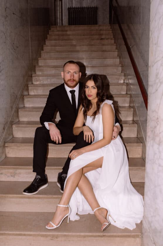 A couple sits on marble stairs; the man wears a black suit and tie, while the woman wears a white dress and heels. They look directly at the camera in a formal indoor setting. by Justin Salem Meyer