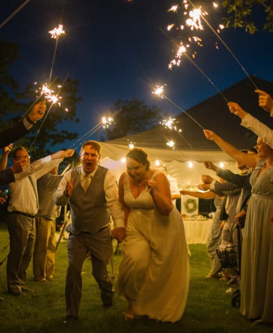A bride and groom laugh and hold hands while running through a tunnel of guests holding sparklers at night during an outdoor wedding celebration. by Justin Salem Meyer