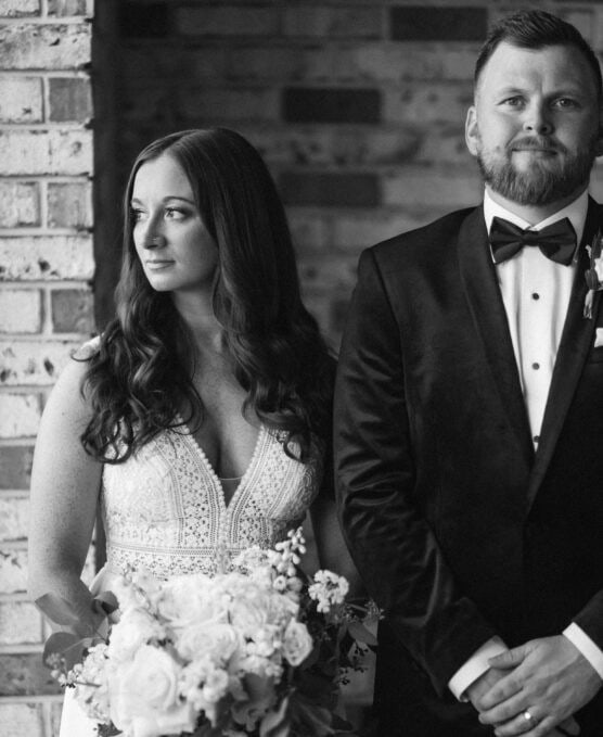 A black and white photo of a bride holding a bouquet and a groom in a tuxedo standing side by side indoors, with a brick wall as the background. The bride looks to the left while the groom faces forward. by Justin Salem Meyer
