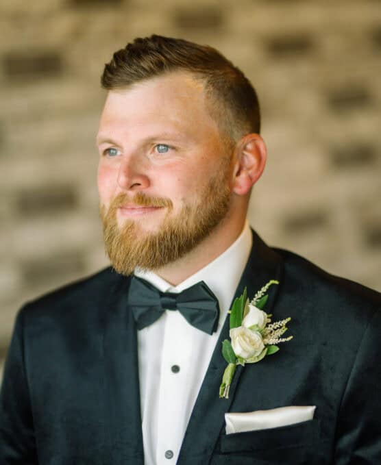 A man with a beard and short hair wears a black tuxedo, white shirt, and black bow tie. He has a white boutonniere pinned to his jacket and is looking slightly to the side, smiling softly, with a blurred background behind him. by Justin Salem Meyer