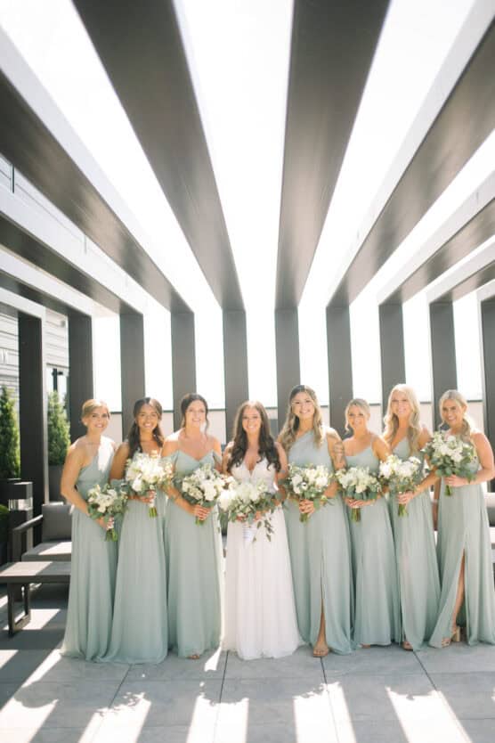 A bride in a white dress stands outdoors with seven bridesmaids in light sage green dresses, all holding bouquets, under modern black and white architectural beams with sunlight streaming through. by Justin Salem Meyer
