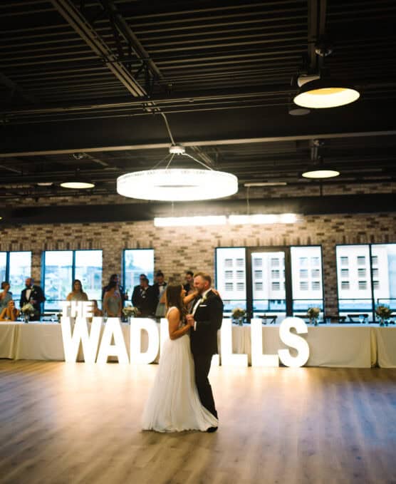 A bride and groom share their first dance in a spacious venue with large windows. Behind them, illuminated letters spell THE WADSWORTHS in front of a long table where guests are seated. by Justin Salem Meyer