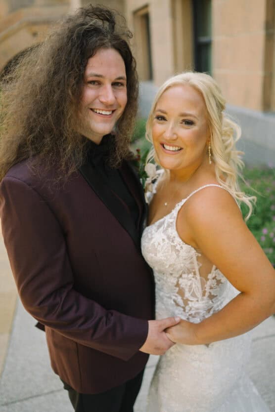 A smiling couple poses closely together outdoors; the man has long curly hair and wears a burgundy suit, while the woman wears a white lace wedding dress with her blonde hair styled in loose curls. by Justin Salem Meyer