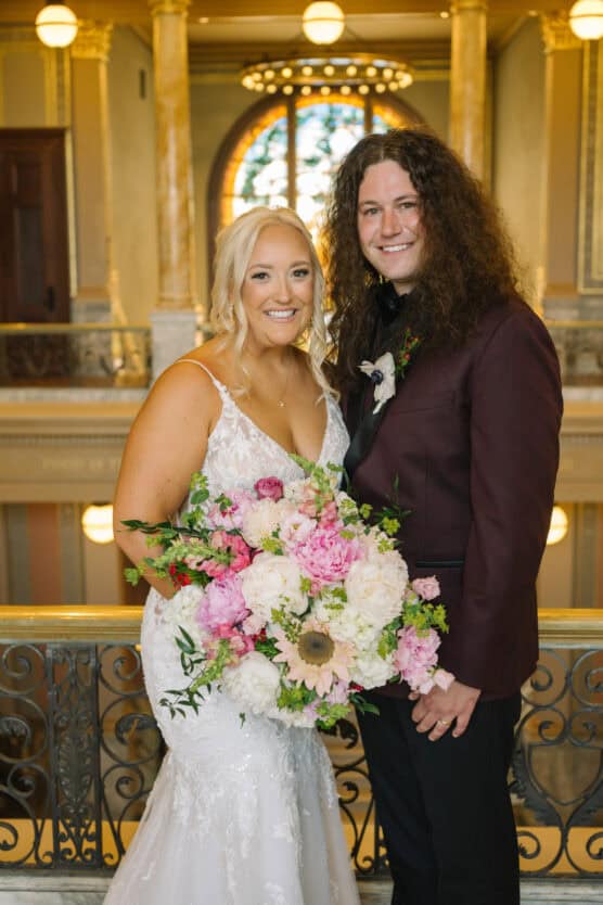 A bride in a white dress holds a large bouquet of pink and white flowers while standing next to a groom in a dark suit. They are smiling indoors with ornate architecture and a stained glass window behind them. by Justin Salem Meyer