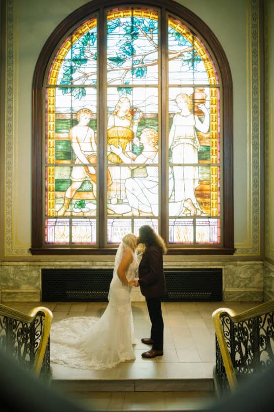 A couple in wedding attire stands holding hands before a large, colorful stained glass window in an ornate building, with intricate railings and marble walls surrounding them. by Justin Salem Meyer