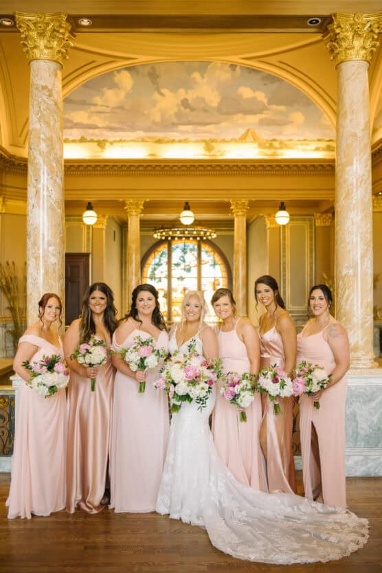 A bride in a white gown stands with six bridesmaids in blush pink dresses, all holding bouquets, in an elegant hall with marble columns and a stained glass window in the background. by Justin Salem Meyer