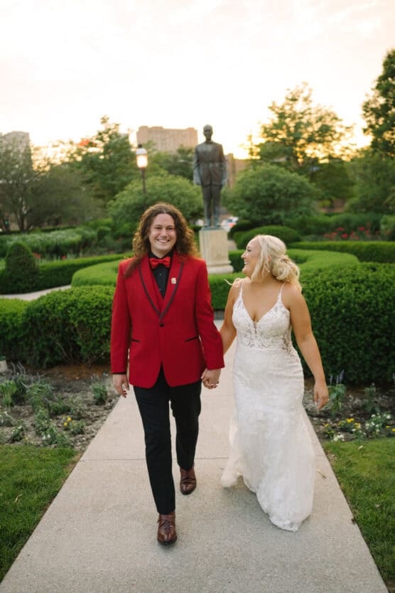 A smiling couple walks hand in hand down a garden path; the man wears a red tuxedo jacket and black pants, and the woman wears a white wedding dress. Green hedges and a statue are visible in the background at sunset. by Justin Salem Meyer