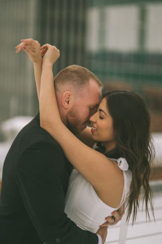 A couple embraces closely, with the womans arms wrapped around the mans neck. They are smiling softly, their faces touching in an intimate moment, set against an urban background. by Justin Salem Meyer