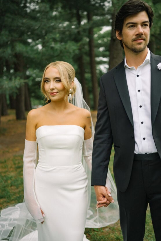A bride in a white off-shoulder gown and veil holds hands with a groom in a black suit and white shirt. They are standing outdoors surrounded by tall trees, looking slightly away from the camera. by Justin Salem Meyer