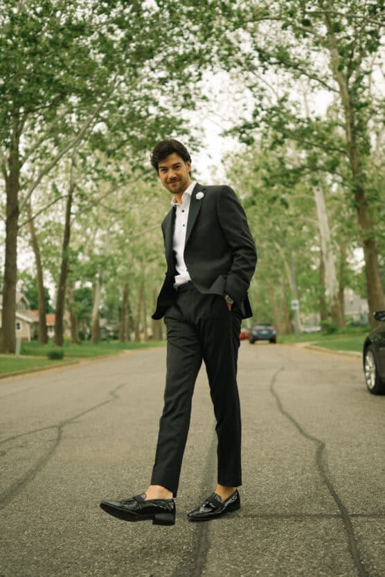 A man in a black suit and dress shoes walks confidently down a tree-lined street, with hands in his pockets and a slight smile, surrounded by greenery and parked cars. by Justin Salem Meyer