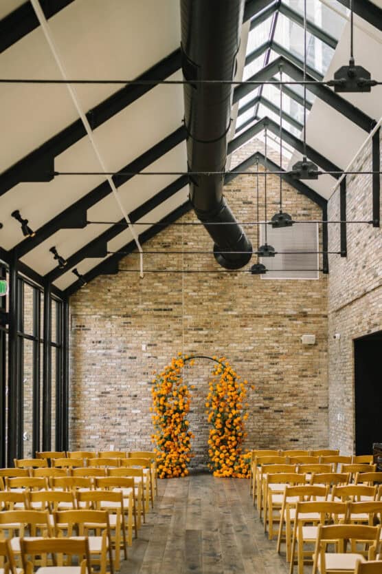 A modern indoor ceremony space with rows of yellow chairs facing a brick wall. An arch made of vibrant orange flowers stands at the front beneath a high, pitched ceiling with exposed beams and ductwork. by Justin Salem Meyer