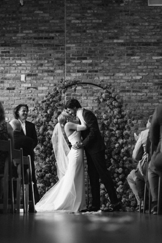A bride and groom share their first kiss at the altar during a wedding ceremony, surrounded by guests. They stand in front of a floral arch and a brick wall, with the scene captured in black and white. by Justin Salem Meyer