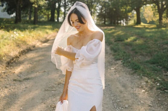 A bride in an off-the-shoulder white dress and veil stands on a sunlit dirt path surrounded by greenery, smiling and looking down. by Justin Salem Meyer