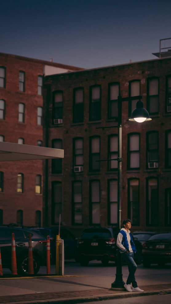 A person in a blue and white jacket stands under a streetlamp on a city sidewalk at dusk, with parked cars and a brick building in the background. by Justin Salem Meyer