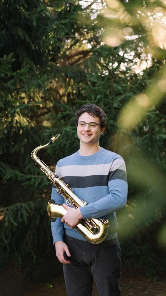 A young person with short curly hair and glasses smiles while holding a saxophone, standing outdoors in front of lush green pine trees. by Justin Salem Meyer