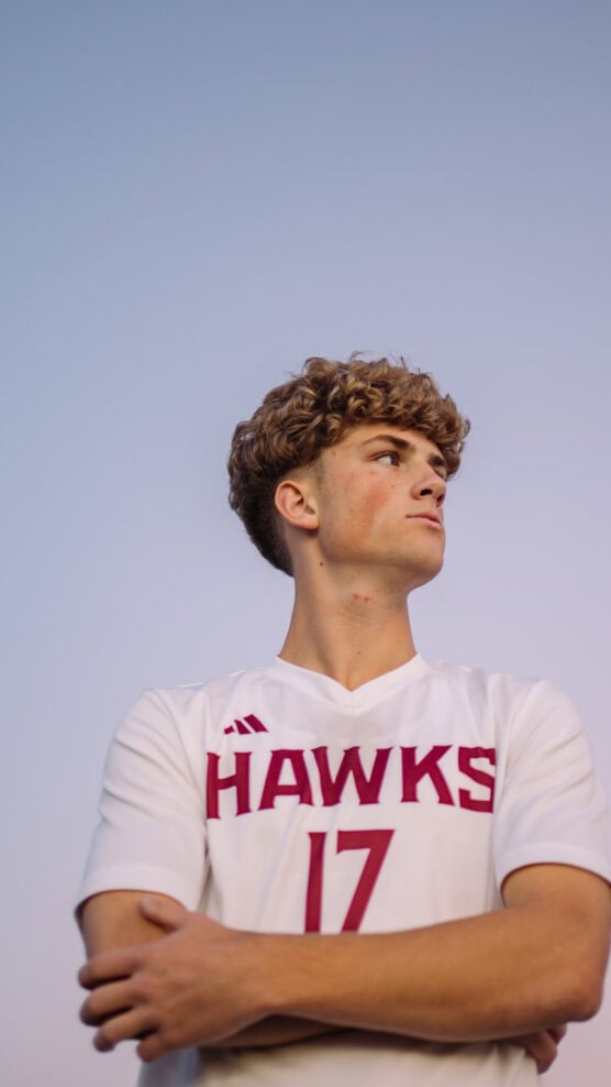 A young man with curly hair wearing a white HAWKS soccer jersey with the number 17 stands confidently with arms crossed, gazing to the side against a clear sky background. by Justin Salem Meyer