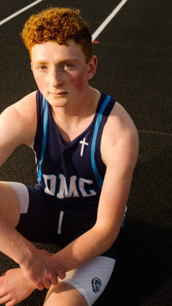 A young athlete with curly red hair sits on a running track, wearing a navy blue and white athletic uniform with the letters OMC and a cross symbol on the chest, looking up at the camera in sunlight. by Justin Salem Meyer