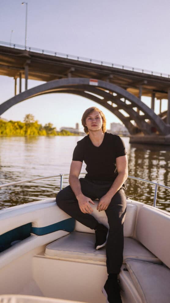 A young man in black clothes sits at the front of a boat on calm water, with a large arched bridge and greenery in the background under a clear sky. by Justin Salem Meyer