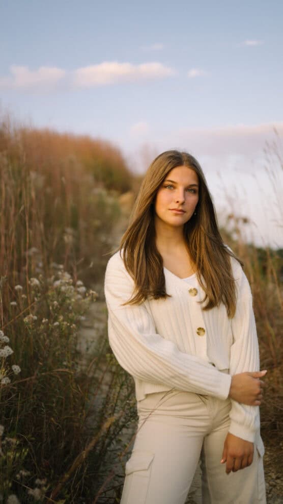 A young woman with long brown hair, wearing a white sweater and light pants, stands in a field of tall grass and wildflowers at sunset, with a calm expression and soft natural light. by Justin Salem Meyer