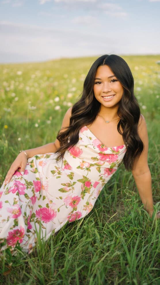 A young woman with long dark hair, wearing a white floral dress, sits smiling in a grassy field with wildflowers under a blue sky. by Justin Salem Meyer