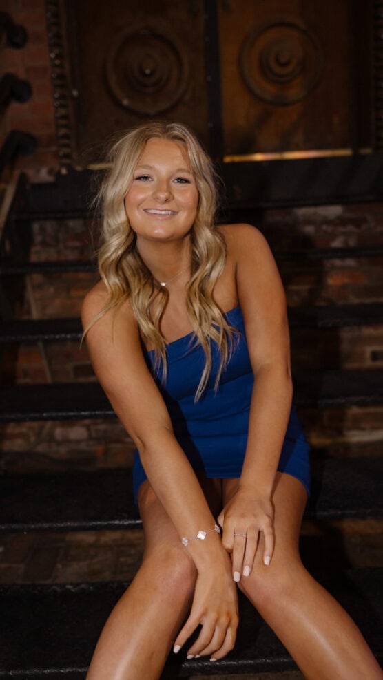 A young woman with long blonde hair wearing a blue dress sits on dark steps, smiling at the camera. There is a brick wall and a large wooden door with metal details in the background. by Justin Salem Meyer
