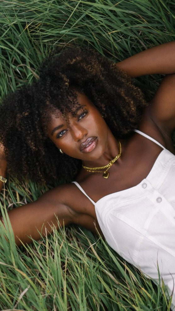 A woman with curly hair lies in tall green grass, wearing a white sleeveless top and gold jewelry, with her arms resting above her head and looking directly at the camera. by Justin Salem Meyer