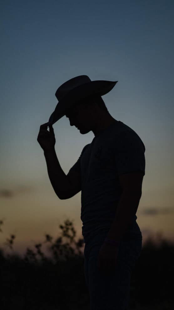 Silhouette of a person wearing a cowboy hat, tilting the brim downward at sunset, with a dusky sky and faint clouds in the background. by Justin Salem Meyer