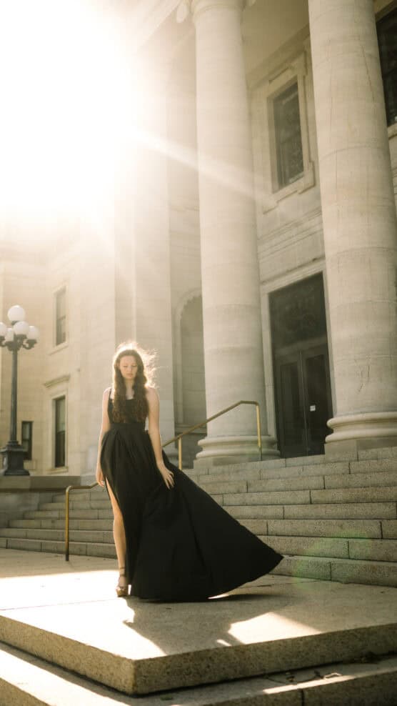 A woman in a long black dress stands on stone steps in front of a grand building with tall columns, as sunlight streams brightly from the top left corner of the image. by Justin Salem Meyer