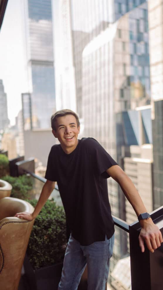 A young person in a black t-shirt and jeans smiles while leaning on a balcony railing, with tall city buildings and skyscrapers in the background. by Justin Salem Meyer
