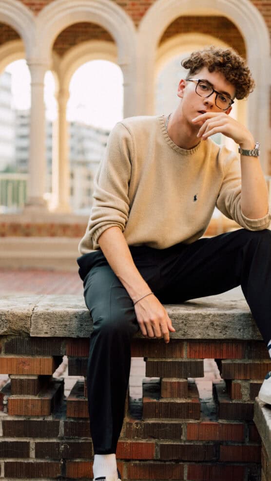 A young man with curly hair and glasses sits on a brick ledge, wearing a beige sweater, black pants, and white socks. He rests one hand on his knee and the other near his face, with arches and buildings in the background. by Justin Salem Meyer