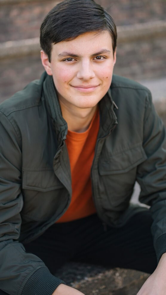 A young person with short dark hair, wearing an olive green jacket over an orange shirt, sits outdoors on stone steps, looking at the camera and smiling gently. by Justin Salem Meyer