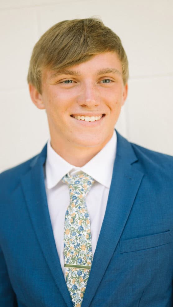 Young man with light brown hair wearing a blue suit, white shirt, and a floral patterned tie, smiling in front of a plain light-colored background. by Justin Salem Meyer