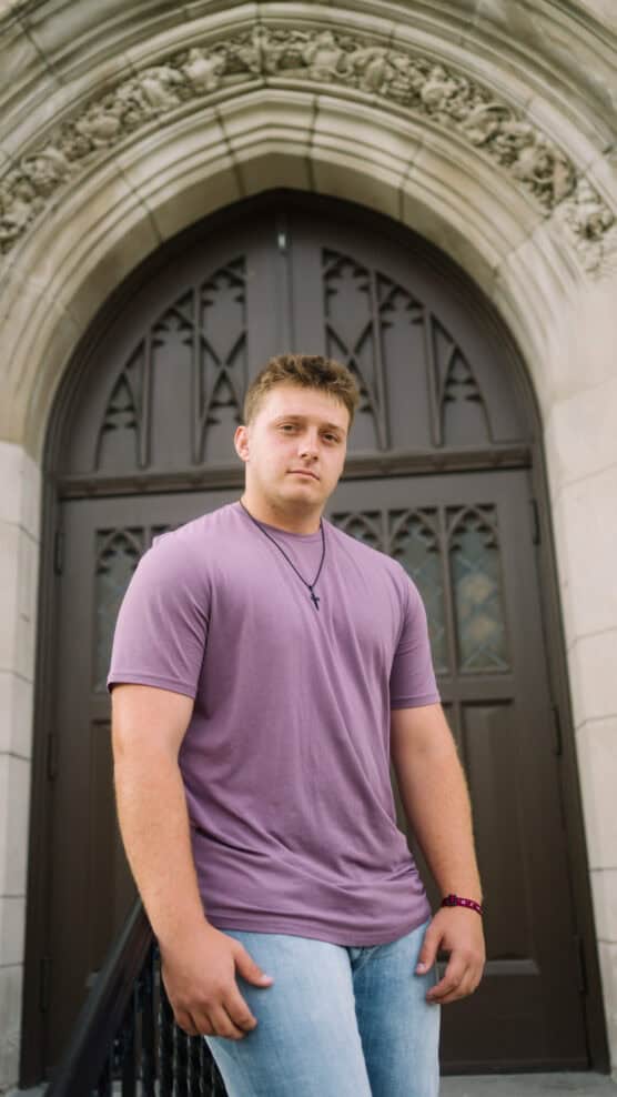 A young man in a purple t-shirt and jeans stands in front of an ornate arched doorway with carved stone details, looking at the camera with a neutral expression. by Justin Salem Meyer