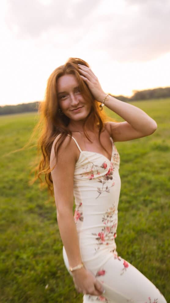 A woman with long red hair wearing a white floral dress smiles and poses outdoors in a green field at sunset, with one hand touching her head and the other by her side. by Justin Salem Meyer