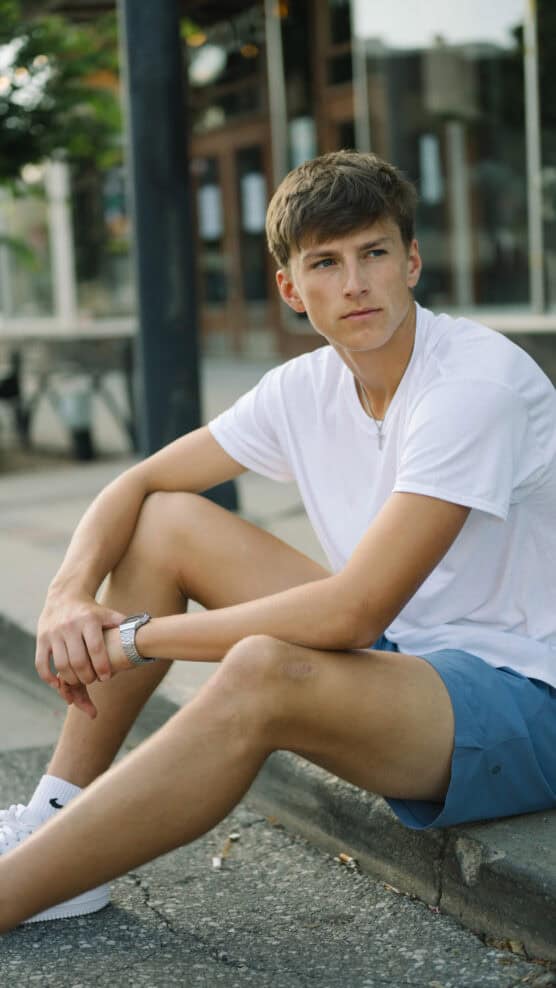 A young person with short brown hair, wearing a white t-shirt, blue shorts, and white sneakers, sits on a curb outside, looking thoughtfully to the side. by Justin Salem Meyer