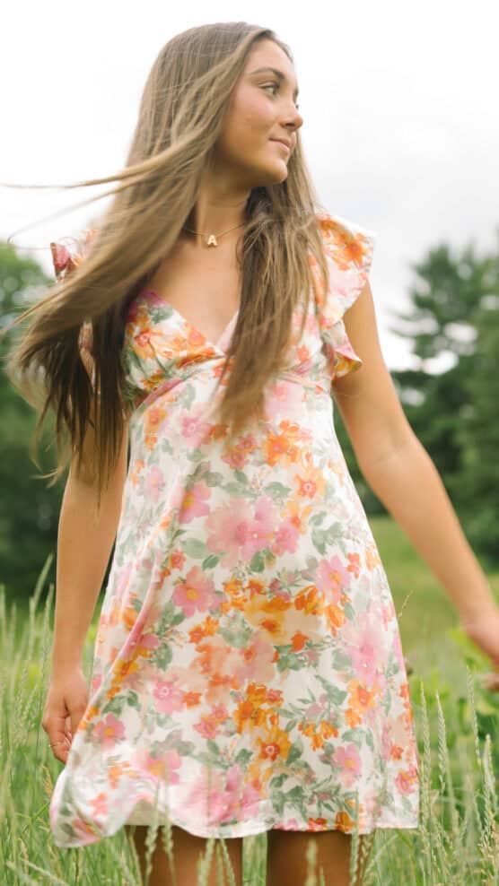 A young woman with long brown hair wearing a colorful floral dress stands in a grassy field, looking to the side with trees and a cloudy sky in the background. by Justin Salem Meyer