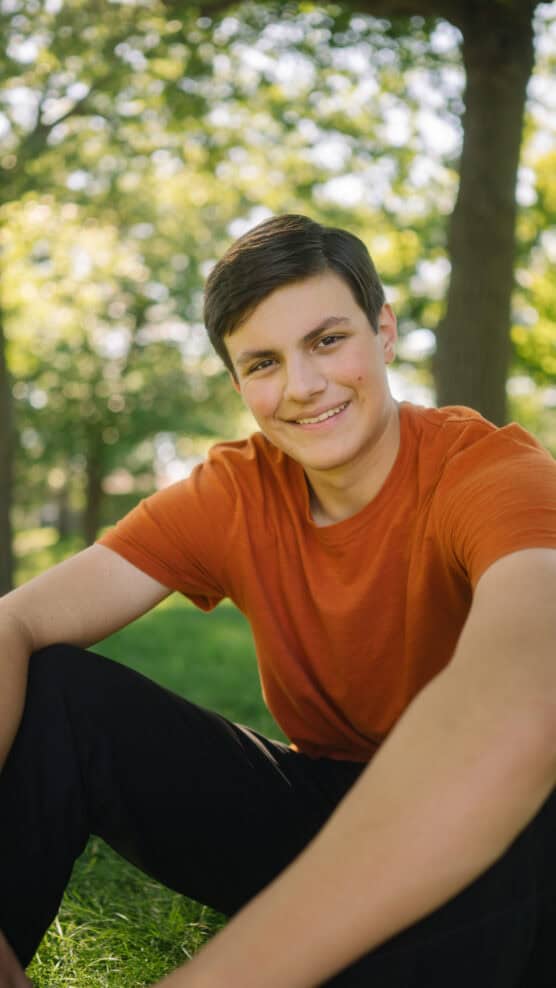 A teenage boy wearing an orange t-shirt sits on grass in a park, smiling at the camera. Sunlight filters through the trees in the background, creating a warm and cheerful atmosphere. by Justin Salem Meyer