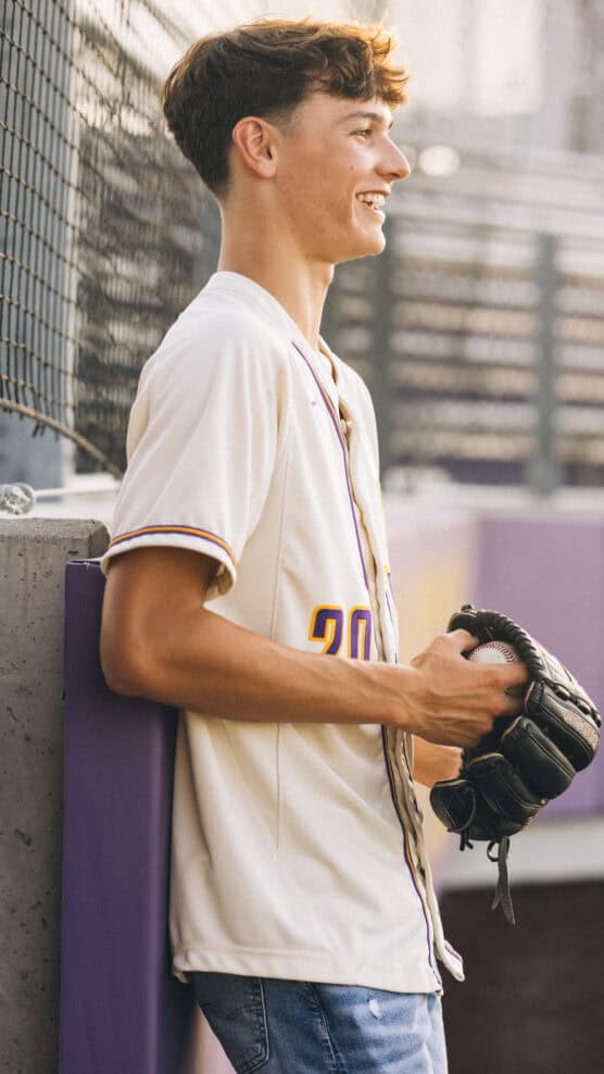 A teenage boy in a white baseball jersey and jeans stands by a fence, smiling and holding a baseball glove, with sunlight illuminating his face. by Justin Salem Meyer