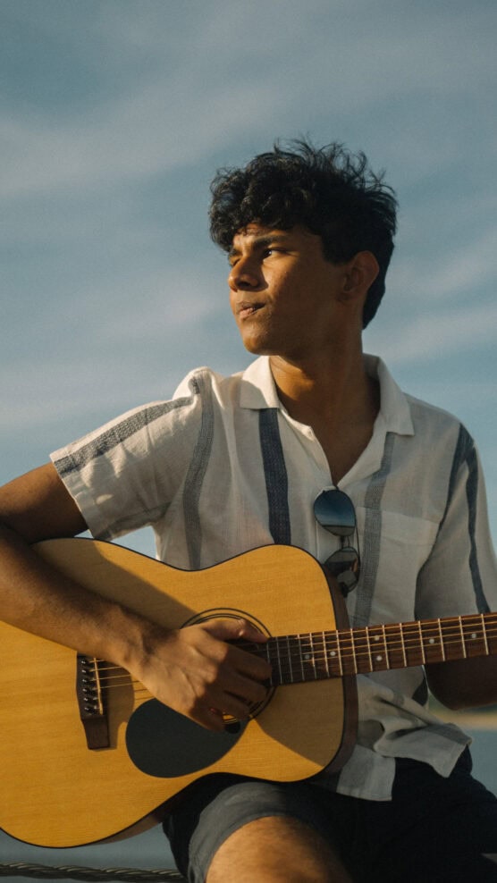 A young man wearing a white short-sleeve shirt and sunglasses, sits outdoors playing an acoustic guitar under a blue sky, looking thoughtfully into the distance. by Justin Salem Meyer