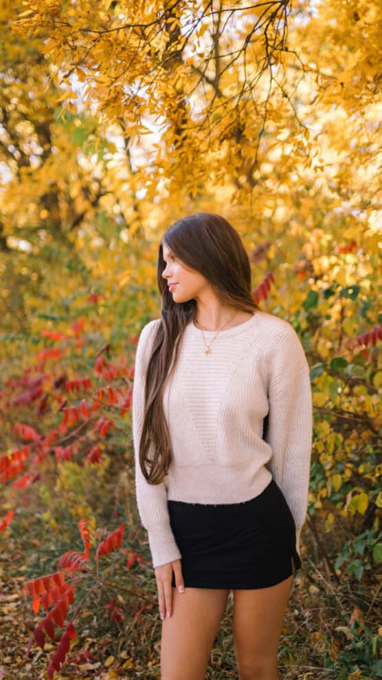 A young woman with long brown hair, wearing a cream sweater and black skirt, stands outdoors among vibrant autumn foliage with yellow and red leaves, looking to her left. by Justin Salem Meyer