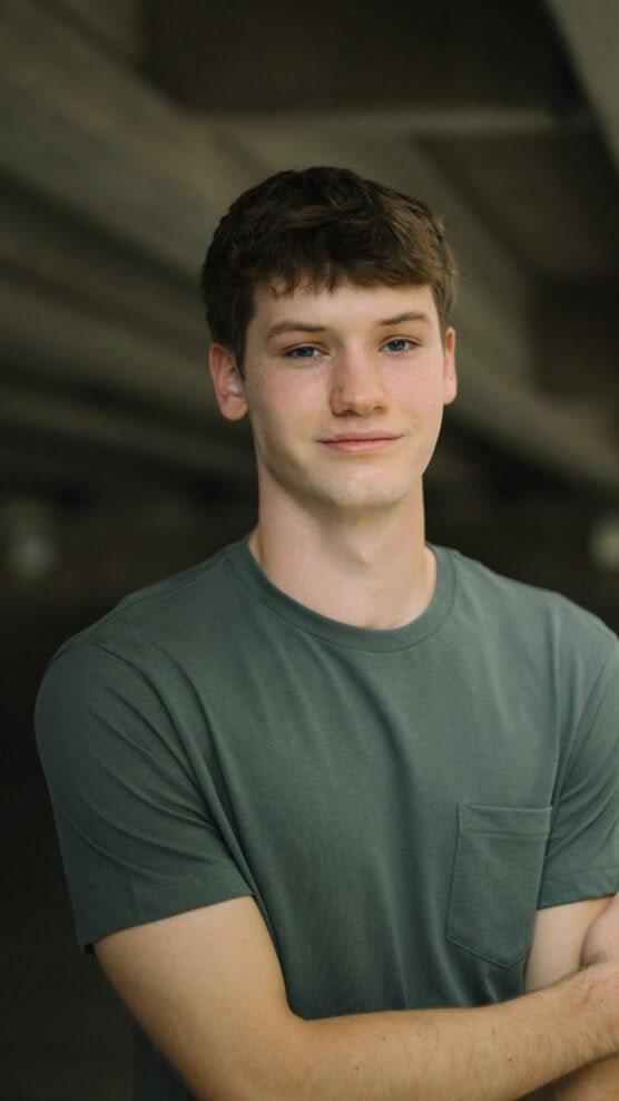 A young man with short brown hair and blue eyes stands smiling slightly, wearing a green t-shirt with a pocket, in front of a blurred, indoor background. by Justin Salem Meyer