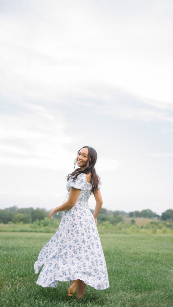 A woman in a light blue floral dress smiles as she twirls in a grassy field under a bright, partly cloudy sky. Trees are visible in the distance, creating a peaceful, summery scene. by Justin Salem Meyer