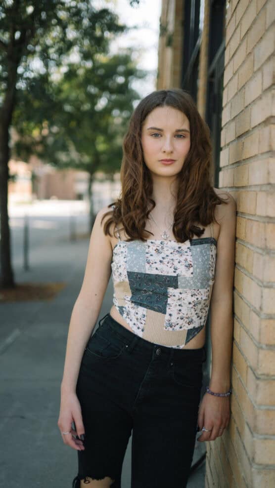 A young woman with wavy brown hair, wearing a patchwork tube top and black jeans, stands leaning against a brick wall on a sidewalk with trees and buildings in the background. by Justin Salem Meyer
